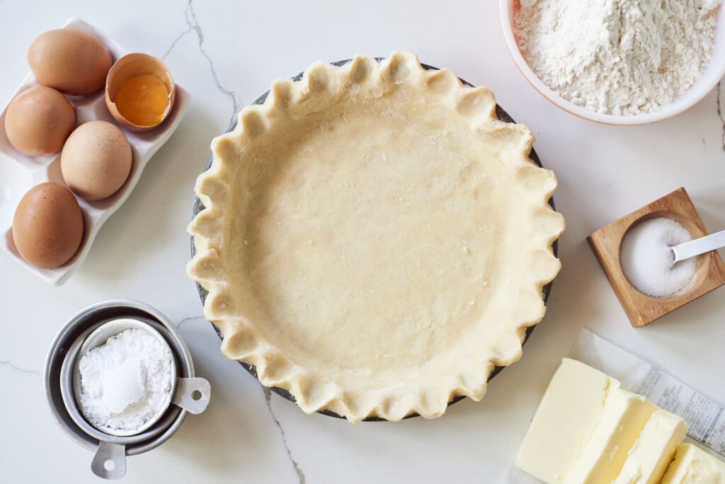 The Perfect Homemade Pie Crust Recipe is assembled in a round pie pan with fluted edges and a buttery, smooth surface. There ingredients including eggs, powdered sugar, all purpose flour and butter around the pie dish.