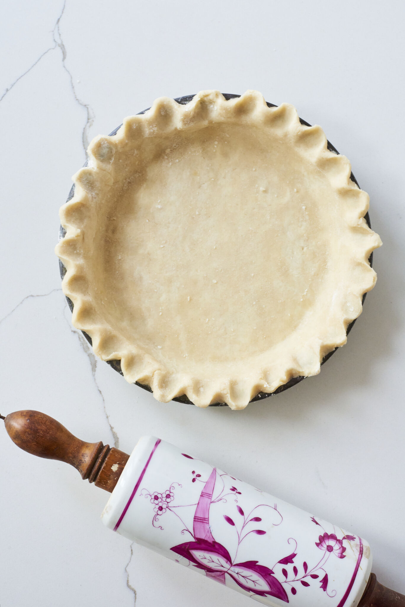A Perfect Homemade Pie Crust with fluted edge is in a round pie pan, with a floral rolling pin on the side. 