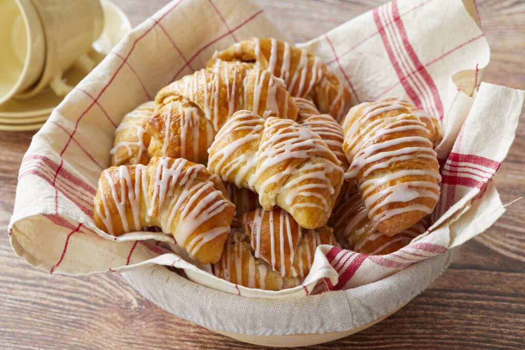 Apple Pie Crescent Rolls are baked golden brown, looking buttery, pillowy, and tender. They're drizzled with vanilla glaze and placed in a linen towel lined proofing basket.