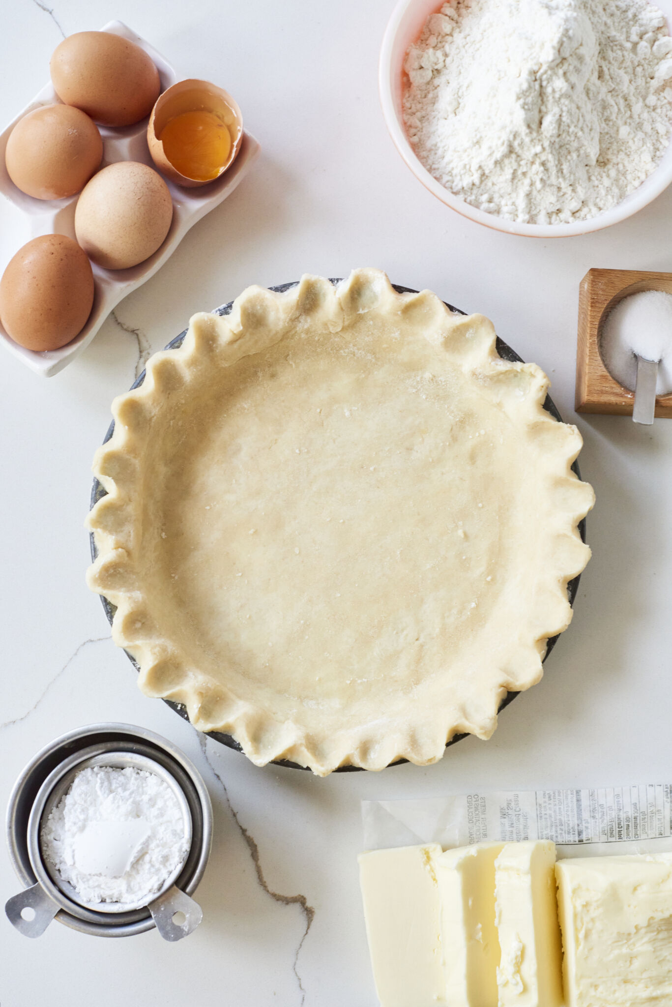 The Perfect Homemade Pie Crust Recipe is assembled in a round pie pan with fluted edges and a buttery, smooth surface. There ingredients including all purpose flour, eggs, powdered sugar, salt, and butter around the pie dish. 