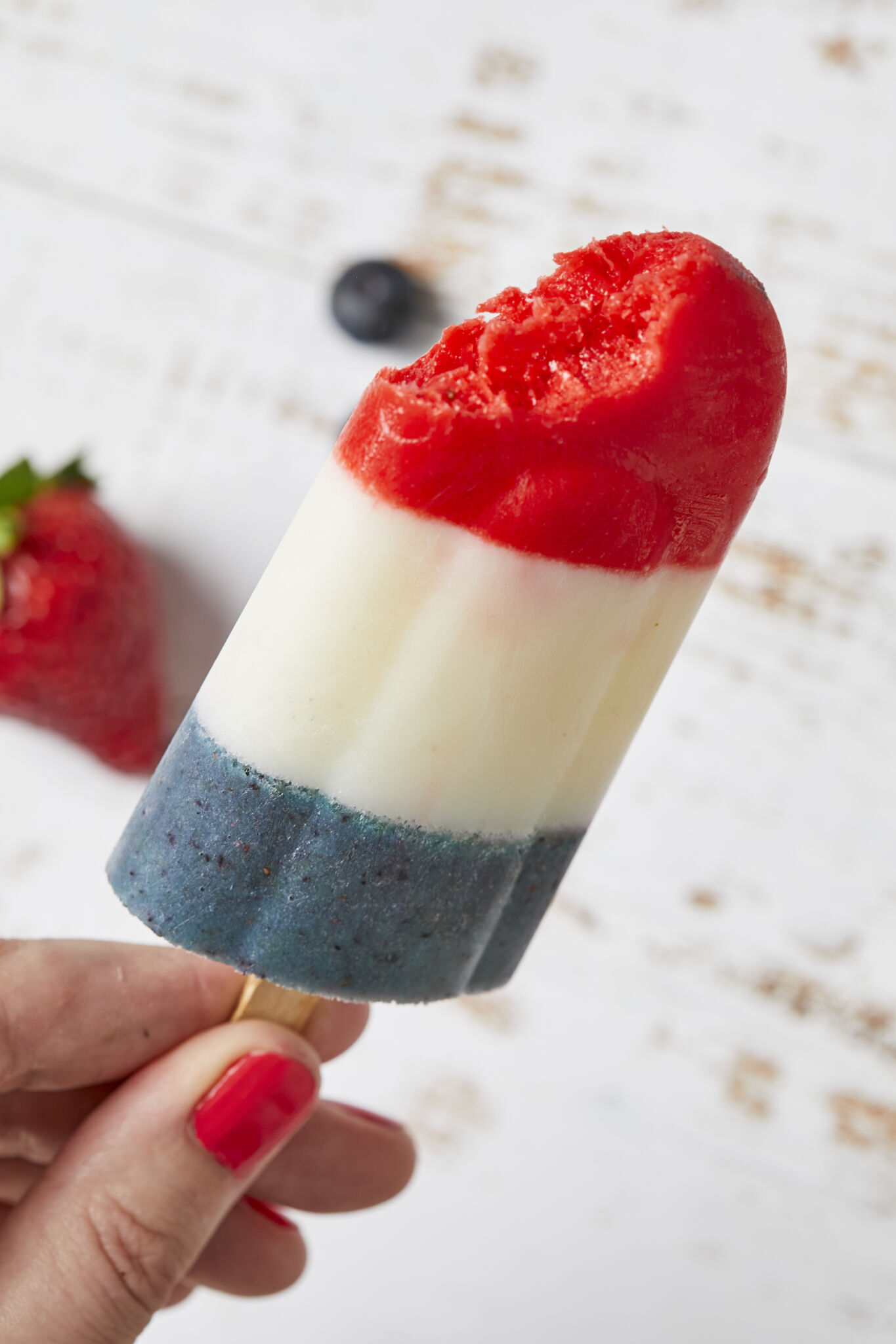 One bomb pop popsicle is being held with fingers by the stick with one bite taken from the red strawberry part on top. 