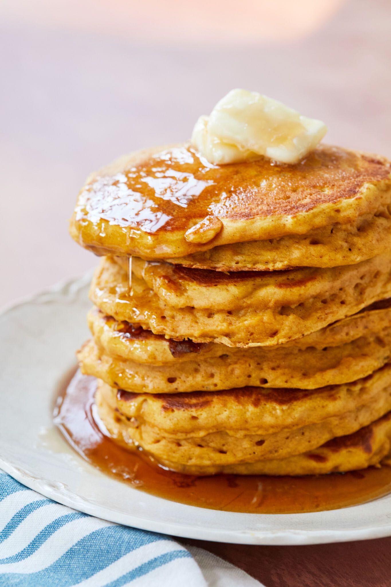 A stack of golden orange Pumpkin Buttermilk Pancakes look fluffy with air bubbles inside, served with maple syrup drizzling from the top on to the plate and crowned with butter. 