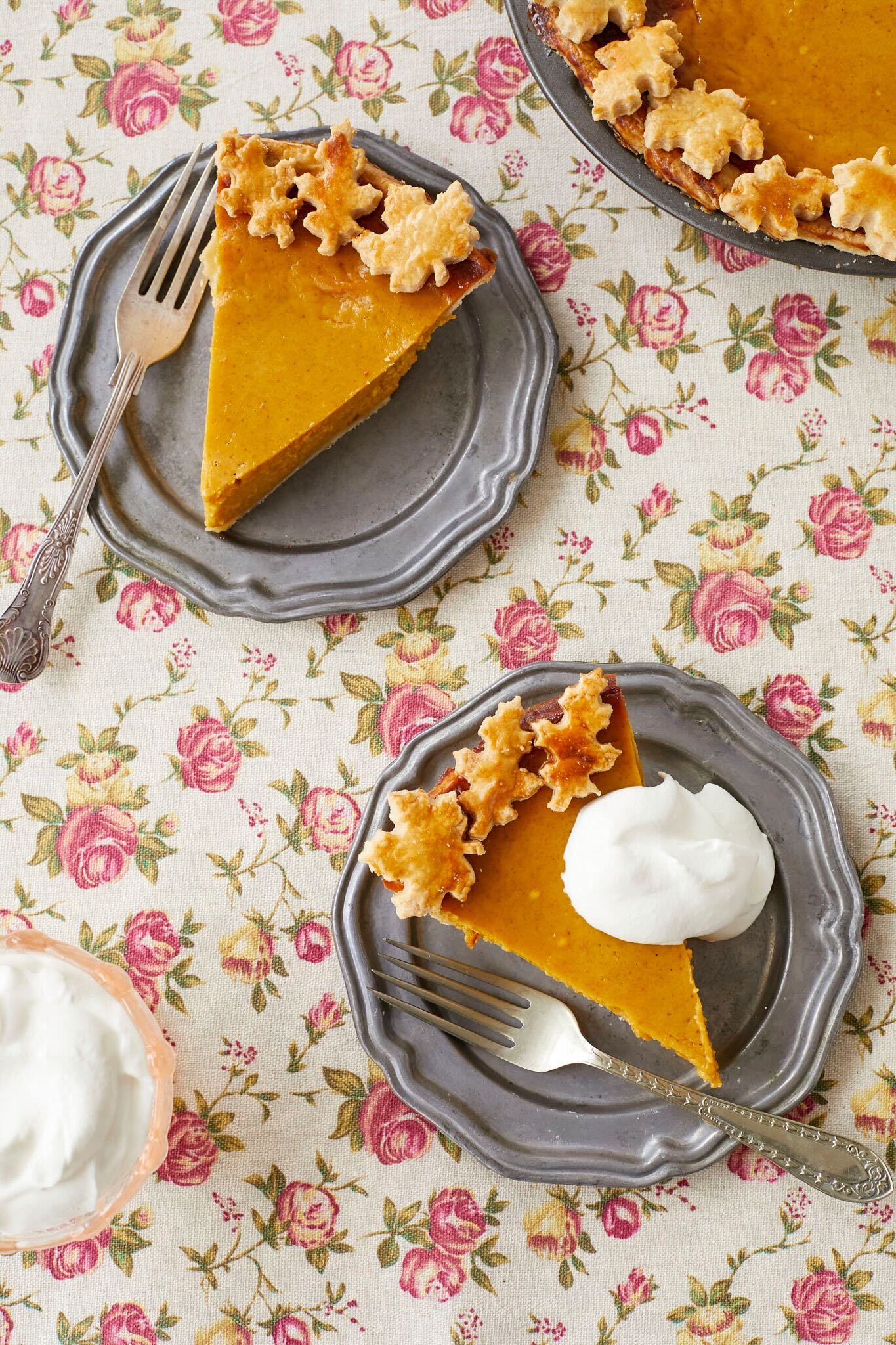 Two slices of homemade pumpkin pie are served on silver dishes. The pie is decorated with leaf-shaped pie crust cut outs and one slice of the pie is topped with a dollop of homemade whipped cream.