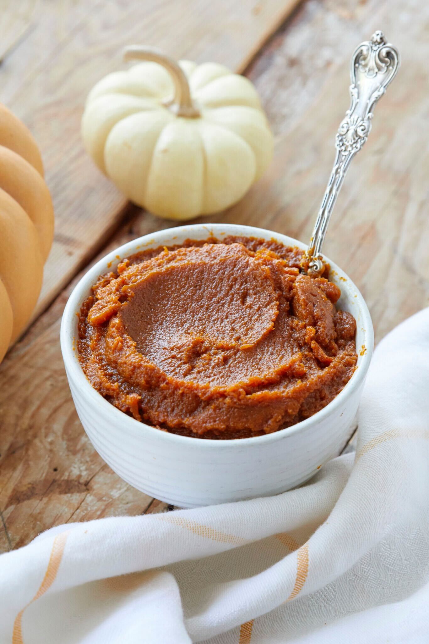 The Coziest Homemade Pumpkin Butter is thick and has a deep orange color. It's served in a small white ceramic bowl with a spoon. 