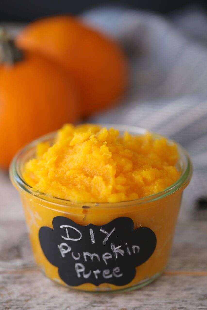 Homemade Pumpkin Puree is served in a glass jar with a vibrant orange-yellow color and smooth consistency.m Two pumpkins are in the background. 