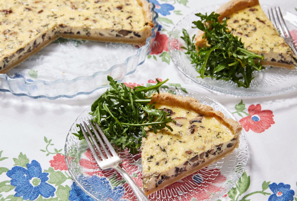 Two slices of Mushroom and Thyme Leaf Tart are cut from the whole tart and served on clear glass plates with forks. The tart has golden, buttery and crumbly crust loaded with velvety custard filling and mushrooms, paired with green earthy thyme leaves. The rest of the tart is in the tart dish.