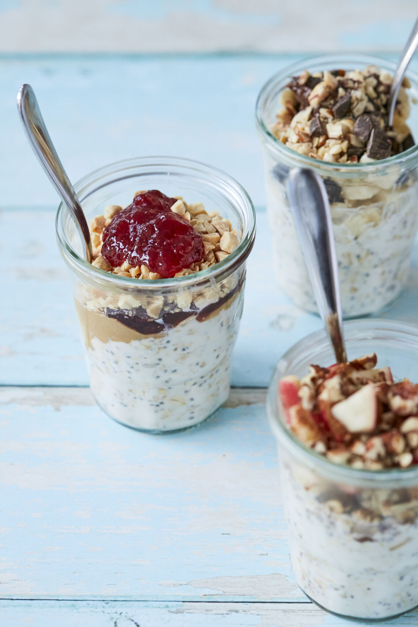 A close-up of 3 jars of Overnight Oats in all 3 flavors with metal spoons in each jar, on a blue wooden table.