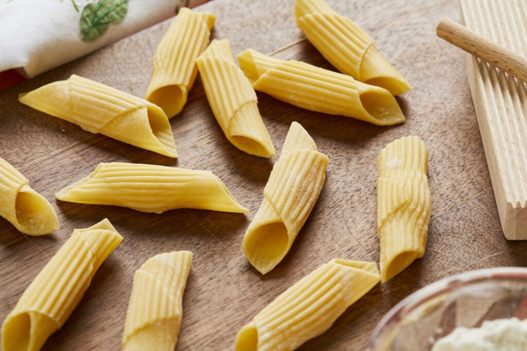 Penne pasta is drying on a wooden board. It's an eggless tubular-shaped pasta, cut diagonally on the ends and resembles an old-fashioned writing quill that was dipped in ink. A small glass bowl of flour is in the bottom right corner.